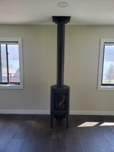 Black wood stove with vertical flue pipe in a bright living room, featuring large windows and dark hardwood flooring.